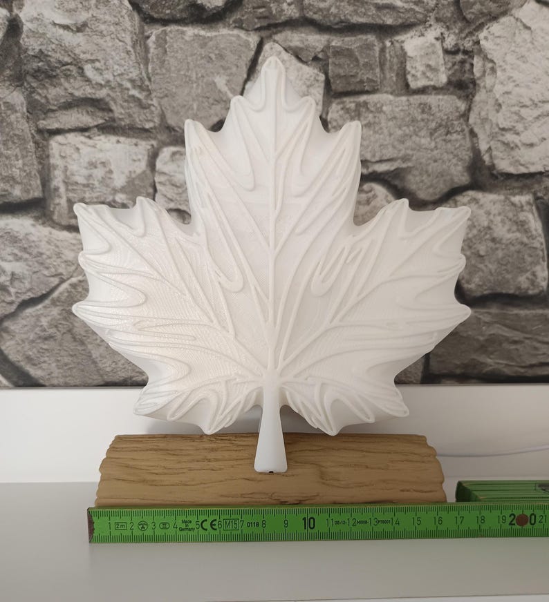 May include: A white maple leaf-shaped object with raised veins, mounted on a textured tan base resembling a log. A green measuring tool is positioned in front, showing the base width is approximately 20 centimeters or 7.87 inches long.