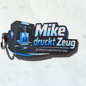 May include: A black and blue keychain featuring a 3D printer graphic and the text Mike druckt Zeug 3D DRUCK UND GESTALTUNG. The design includes a stylized letter M on the printer bed, with a metal ring attached to the left side of the flat object.