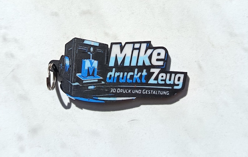 May include: A black and blue keychain featuring a 3D printer graphic and the text Mike druckt Zeug 3D DRUCK UND GESTALTUNG. The design includes a stylized letter M on the printer bed, with a metal ring attached to the left side of the flat object.