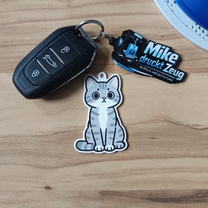 May include: A gray and white tabby cat keychain with large black eyes, attached to a black car key fob and a black rectangular tag featuring the white text Mike druckt Zeug and 3D DRUCK UND GESTALTUNG with a blue 3D printer icon.