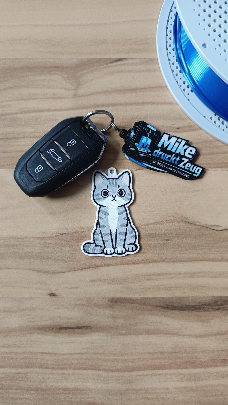 May include: A gray and white tabby cat keychain with large black eyes, attached to a black car key fob and a black rectangular tag featuring the white text Mike druckt Zeug and 3D DRUCK UND GESTALTUNG with a blue 3D printer icon.