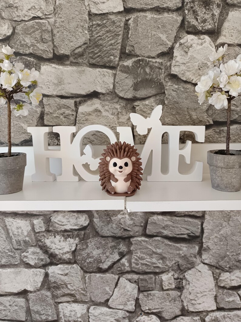 以下が含まれることがあります： 白い「HOME」のサイン、小さなハリネズミの置物、2つの鉢植えの植物が特徴の装飾的な棚の配置。背景は石模様の壁です。