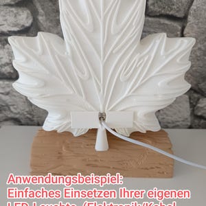 May include: A white 3D-printed maple leaf ornament with a central hole, mounted on a textured tan base resembling a wooden log. A white cable is attached to the back of the leaf. The text on the image reads: Anwendungsbeispiel: Einfaches Einsetzen Ihrer eigenen LED-Leuchte. (Elektronik/Kabel NICHT im Lieferumfang enthalten)
