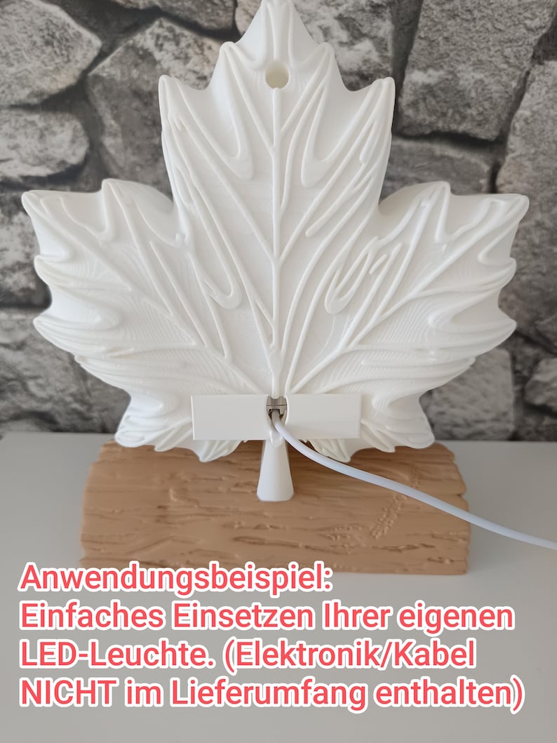 May include: A white 3D-printed maple leaf ornament with a central hole, mounted on a textured tan base resembling a wooden log. A white cable is attached to the back of the leaf. The text on the image reads: Anwendungsbeispiel: Einfaches Einsetzen Ihrer eigenen LED-Leuchte. (Elektronik/Kabel NICHT im Lieferumfang enthalten)