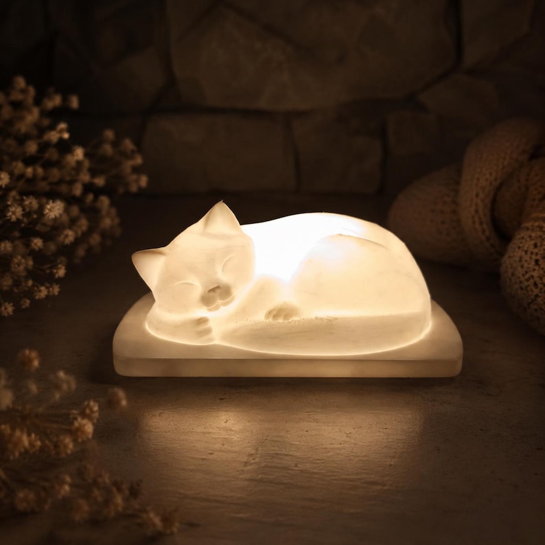 K&ouml;nnte beinhalten: Eine wei&szlig;e, durchscheinende, leuchtende Lampe in Form einer schlafenden Katze auf einem rechteckigen Sockel. Das Licht strahlt aus der Katzenfigur heraus, betont ihre glatte, geschwungene Form und die geschlossenen Augen und erzeugt sanftes Licht.