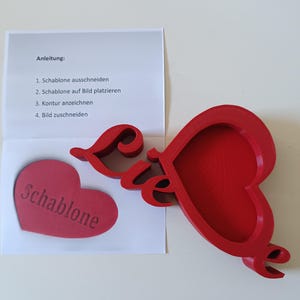 May include: A red heart-shaped craft template with the word "Schablone" and the word "Liebe" in script. A paper with instructions in German is also present. The template is likely for crafting or DIY projects.