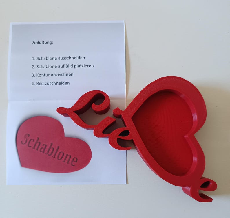 May include: A red heart-shaped craft template with the word "Schablone" and the word "Liebe" in script. A paper with instructions in German is also present. The template is likely for crafting or DIY projects.