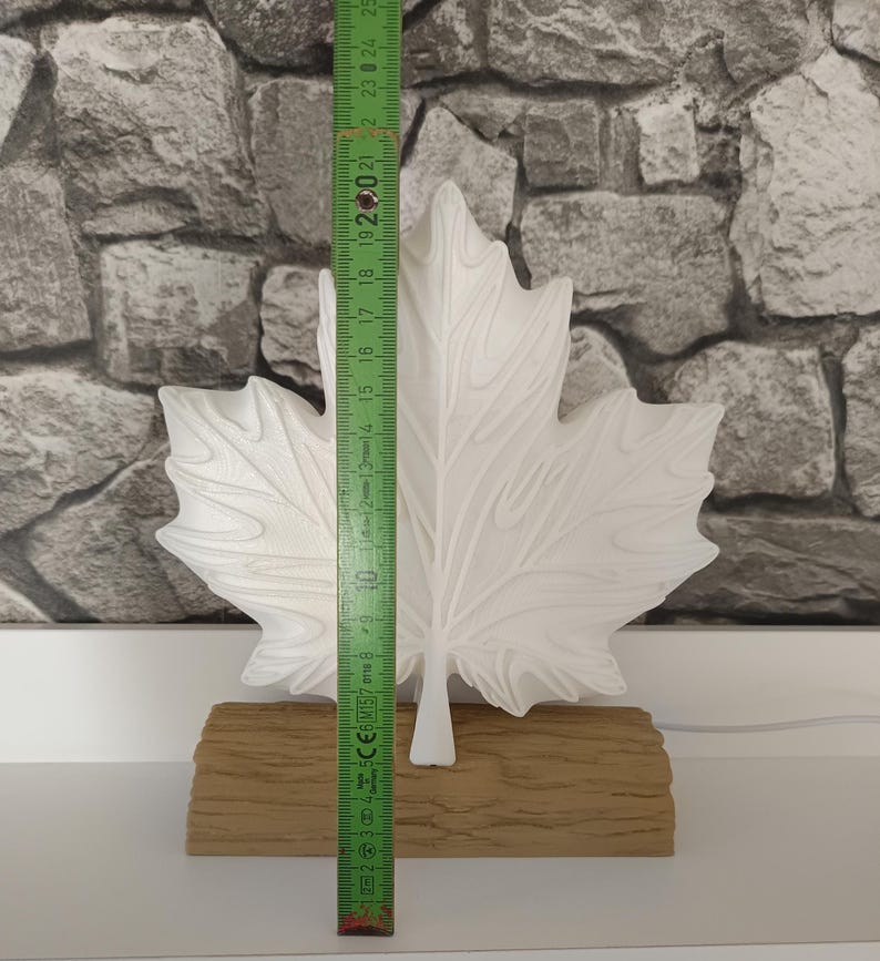 May include: A white maple leaf-shaped decorative object mounted on a textured tan base. A green folding ruler is placed vertically against the leaf, showing a height of approximately 9.8 inches from the base to the top tip of the leaf.
