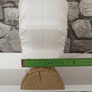 May include: A white lamp with a textured, geometric surface sits on a base shaped like a brown wooden log. A green measuring tape is positioned across the front, showing the base width is approximately 4.75 inches wide at the widest point.