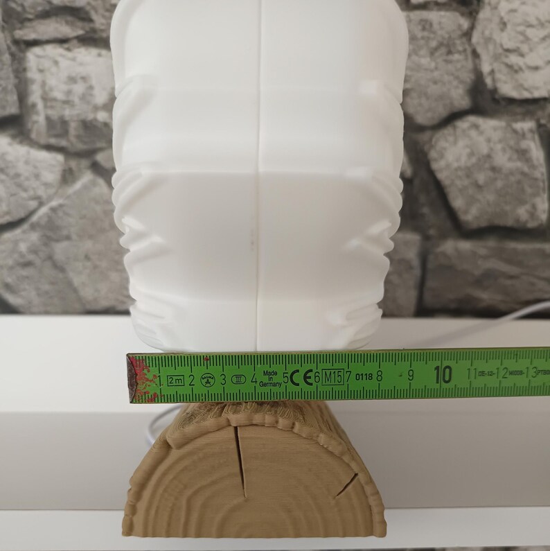 May include: A white lamp with a textured, geometric surface sits on a base shaped like a brown wooden log. A green measuring tape is positioned across the front, showing the base width is approximately 4.75 inches wide at the widest point.