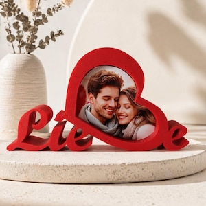 May include: A red heart-shaped photo frame with the word "Liebe" in cursive script. The frame holds a photo of a smiling couple. The frame is on a white surface with a vase and decorative plants in the background.