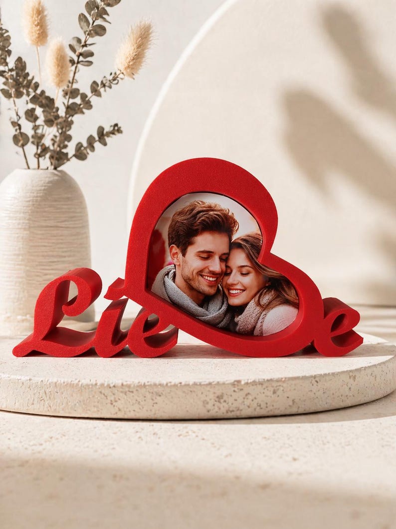 May include: A red heart-shaped photo frame with the word "Liebe" in cursive script. The frame holds a photo of a smiling couple. The frame is on a white surface with a vase and decorative plants in the background.