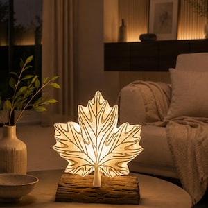 May include: A table lamp shaped like a large maple leaf with a translucent white surface and visible vein details. The leaf is mounted on a textured, brown, log-shaped base. The lamp emits a warm, soft glow from within, illuminating the leaf structure.