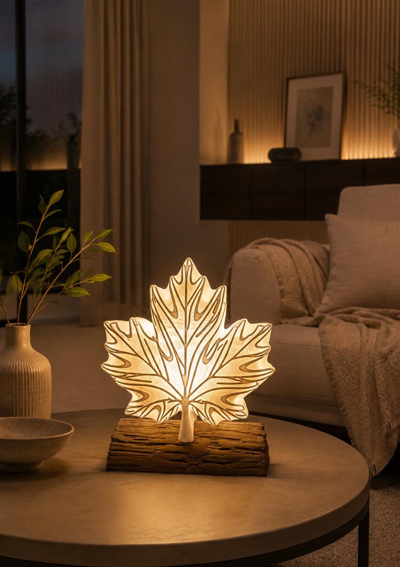 May include: A table lamp shaped like a large maple leaf with a translucent white surface and visible vein details. The leaf is mounted on a textured, brown, log-shaped base. The lamp emits a warm, soft glow from within, illuminating the leaf structure.