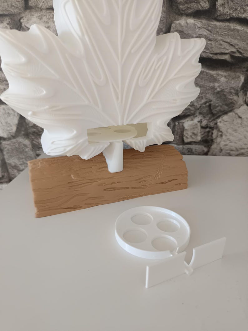 May include: A white 3D-printed maple leaf sculpture mounted on a textured tan log-shaped base. A separate white circular tray with four small indentations and a rectangular connector piece are positioned in front of the main leaf structure.