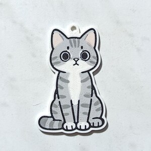 May include: A flat, die-cut acrylic charm featuring a cartoon illustration of a grey tabby cat with dark grey stripes, large black eyes, and a white chest patch. The cat is in a seated position with its tail curled around its side and a small hole at the top for hanging.
