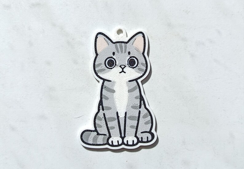 May include: A flat, die-cut acrylic charm featuring a cartoon illustration of a grey tabby cat with dark grey stripes, large black eyes, and a white chest patch. The cat is in a seated position with its tail curled around its side and a small hole at the top for hanging.