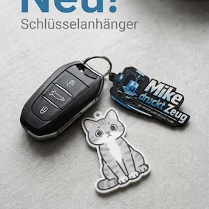 May include: A black car key fob attached to a metal ring with two keychains: one is a grey and white tabby cat illustration, and the other is a black and blue tag with the text Mike druckt Zeug 3D DRUCK UND GESTALTUNG.