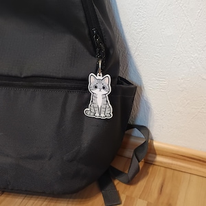 May include: A gray and white tabby cat keychain attached to the zipper of a dark gray backpack. The keychain features a cartoon-style illustration of a sitting cat with large eyes, a small nose, and distinct stripes on its head, body, and tail.