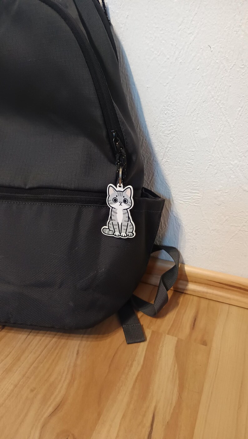 May include: A gray and white tabby cat keychain attached to the zipper of a dark gray backpack. The keychain features a cartoon-style illustration of a sitting cat with large eyes, a small nose, and distinct stripes on its head, body, and tail.