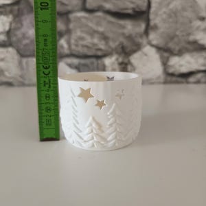 以下が含まれることがあります： 松の木と星の切り抜きデザインが施された白い円筒形のキャンドルホルダー。緑色の測定ツールがオブジェクトの横に垂直に配置されており、キャンドルホルダーの高さが約6.5cmであることを示しています。