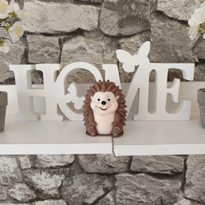 以下が含まれることがあります： 笑顔のハリネズミの装飾品。茶色の針を持ち、白い「HOME」サインと蝶のアクセントの前に配置されています。背景は灰色の石壁です。