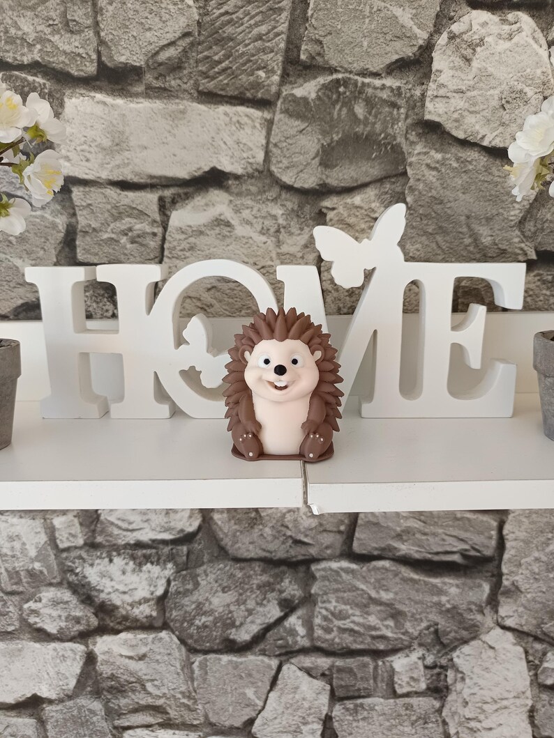 以下が含まれることがあります： 笑顔のハリネズミの装飾品。茶色の針を持ち、白い「HOME」サインと蝶のアクセントの前に配置されています。背景は灰色の石壁です。