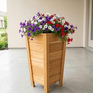 May include: A tall, square wooden planter box with a natural light brown finish and horizontal slat construction. The planter is filled with a dense arrangement of vibrant petunia flowers in shades of deep purple, bright red, and crisp white.