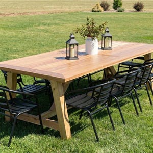 Puede incluir: Una mesa de comedor rectangular de madera marrón claro con base de caballete y ocho sillas de metal negro con listones horizontales. Dos faroles de metal negro y un jarrón blanco con follaje verde se sitúan sobre la mesa.