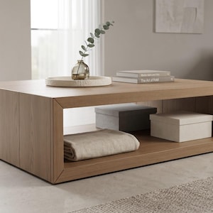 Puede incluir: Mesa de centro rectangular de madera clara con estante inferior. Diseño minimalista. Una manta enrollada, dos cajas, libros y un jarrón decorativo están sobre la mesa. La mesa mide aproximadamente 120 cm de largo.