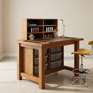 May include: A wooden craft desk with a built-in hutch and storage drawers. The desk has a dark surface and is accompanied by a yellow adjustable stool and a silver desk lamp. The hutch has shelves with art supplies.