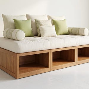 Könnte beinhalten: Ein modernes Tagesbett mit einem hellen Holzsockel mit drei offenen Staufächern. Das Tagesbett ist mit einer cremefarbenen, getufteten Matratze und mehreren Kissen in Creme- und Grüntönen ausgestattet.
