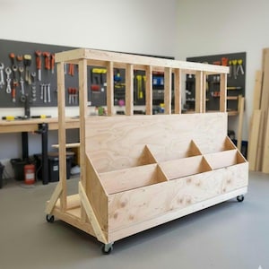 May include: A large, unfinished wooden storage cart with multiple compartments and a slatted top. The cart is on wheels and appears to be designed for workshop or garage use. The wood has a natural, light colour.