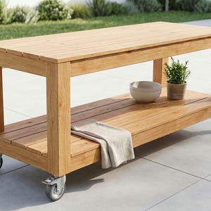 May include: A rectangular light brown wooden kitchen island cart featuring a slatted top and a lower shelf supported by four sturdy legs with silver metal caster wheels. A beige cloth, a small white bowl, and a potted green plant sit on the shelf.