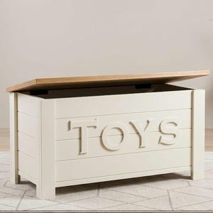 May include: A rectangular off-white wooden toy chest with a natural light wood lid. The front panel features horizontal slats and the word TOYS in large, raised, matching off-white capital letters. The lid is partially open, revealing the interior.