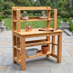 May include: A wooden potting bench with a two-tiered shelf and lower storage shelves. The bench holds a succulent, gardening gloves, a copper watering can, a leather apron, a metal bucket, and a terracotta pot. The bench is outdoors on a stone patio.