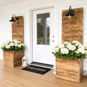 May include: Two tall wooden planters with attached back panels and black wall-mounted lamps, each containing a large cluster of white hydrangea flowers. A black rectangular doormat with the white text WELCOME sits before a white front door.