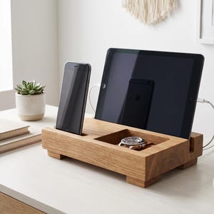Op de afbeelding: Een rechthoekig houten dockingstation met een gleuf voor een smartphone, een bredere gleuf voor een tablet en een vierkant verzonken vak met een zilveren polshorloge. De standaard heeft vier kleine voetjes en uitsparingen aan de zijkanten.