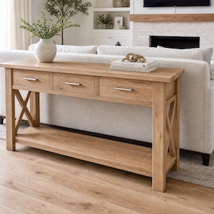 May include: A light oak console table featuring three drawers with silver-toned horizontal handles, an open bottom shelf, and X-shaped side supports. The top surface holds a white vase with greenery and two stacked books with a decorative chain link.