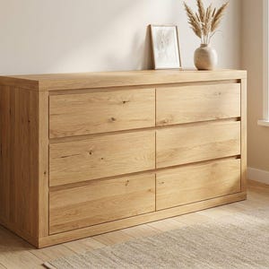 Op de afbeelding: Een licht eikenhouten dressoir met zes laden en een natuurlijke houtnerf afwerking. Het dressoir heeft een rechthoekige vorm en een platte bovenkant. Een ingelijste foto en een vaas met gedroogde planten staan erop. Het dressoir staat in een goed verlichte kamer.