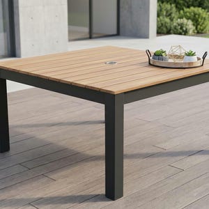 May include: A square outdoor dining table featuring a light brown slatted wood tabletop with a central umbrella hole and a sturdy black metal frame. A round wooden tray with small potted succulents sits on the surface of the table.