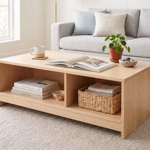 May include: A rectangular light wood coffee table featuring an open shelf design with two storage compartments. The top surface holds an open book, a two-toned ceramic mug, a small potted plant, and a bowl of stones. A woven basket sits below.