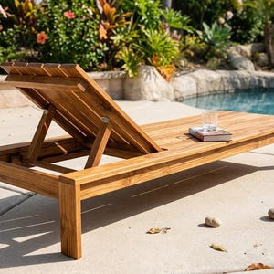May include: A natural brown wooden lounge chair with a slatted surface and an adjustable backrest. The chair features a rectangular frame with four sturdy legs and a flat seat area. A small book and a clear glass are placed on the flat section.