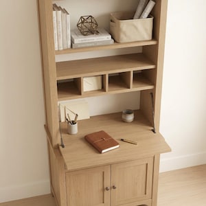 May include: A light oak desk and hutch unit. The desk features a drop-down writing surface and a two-door cupboard. The hutch has shelves with books, a basket, and decorative items. A notebook and pen are on the desk.