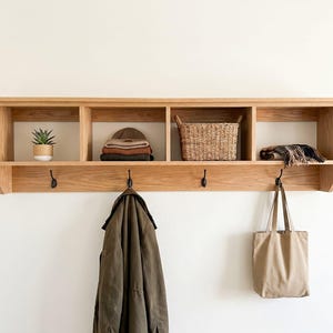 May include: A light-toned wooden wall-mounted storage unit featuring four open cubby compartments and four black metal hooks. The shelves hold a small potted succulent, a stack of folded garments, a woven basket, and a patterned textile item.