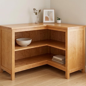 May include: A light brown wooden corner cabinet featuring an L-shaped design with a flat top surface and two open shelves. The unit is constructed with vertical side panels and sturdy legs, providing an open storage area for organising items.