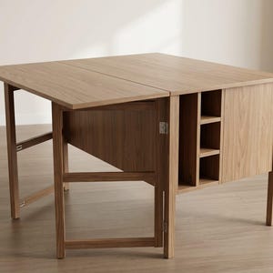 May include: A light brown, wooden drop-leaf table with a rectangular top. The table has a cabinet with shelves on one side and is supported by four legs. The table is 30 inches wide when the leaf is down.