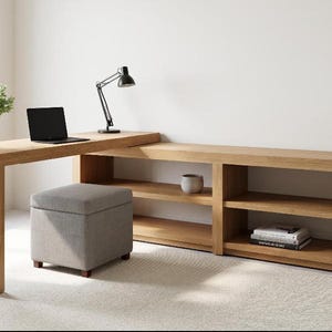 May include: A modern home office setup featuring a light wood desk with a built-in bookshelf. A black laptop and desk lamp sit on the desk. A gray ottoman and a white vase add to the decor. The desk is approximately 183 cm long.