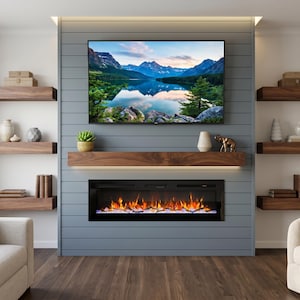 Könnte beinhalten: Ein wandmontierter elektrischer Kamin mit schwarzem Glasrahmen und leuchtend orangefarbenen Flammen unter einem Flachbildfernseher, der eine Bergseelandschaft zeigt, vor einer grauen Wand mit horizontalen Paneelen und Holzregalen.
