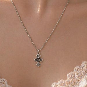 May include: A silver-colored necklace with a small, ornate cross pendant. The cross features intricate detailing and is suspended from a delicate chain. The necklace is worn against a light-colored background.